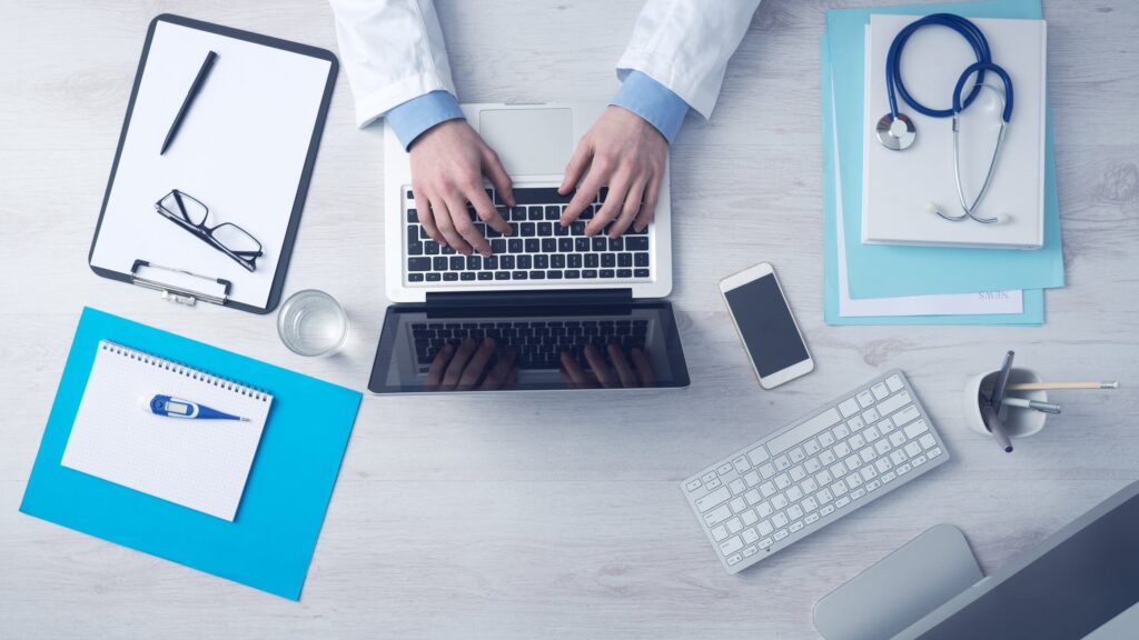 Médico digitando em notebook em mesa de trabalho com estetoscópio, prancheta e materiais de escritório, representando planejamento de publicidade médica ética.